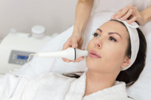 Adult woman enjoying in face massage after hyaluron injection in lips in wellness center. Portrait of beautiful female in ordination.